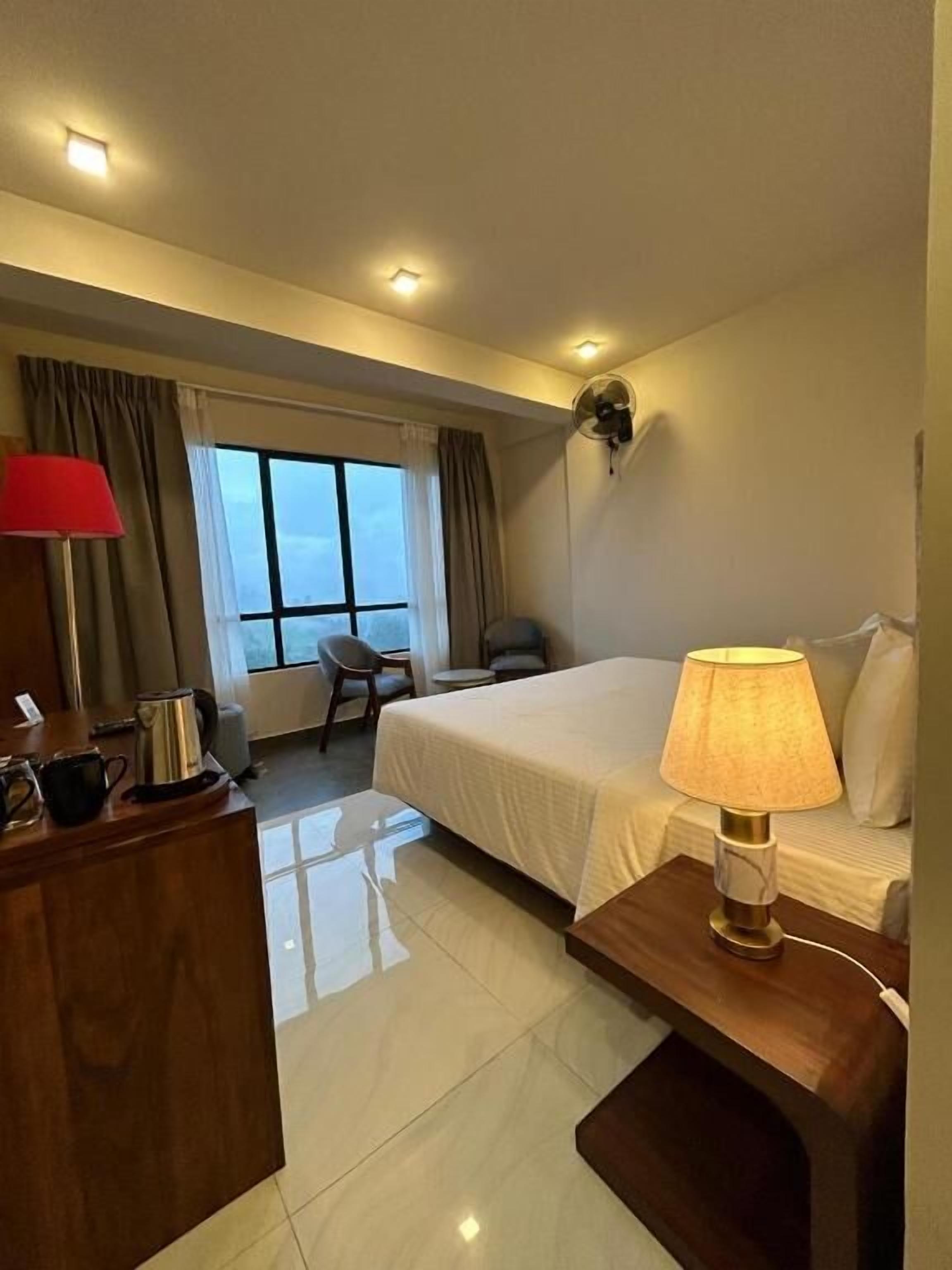 Ein Hotelzimmer mit Bett, Nachttisch, Lampe, Schreibtisch mit Kaffeemaschine, Stuhl und einem Vorhang drapierten Fenster.
