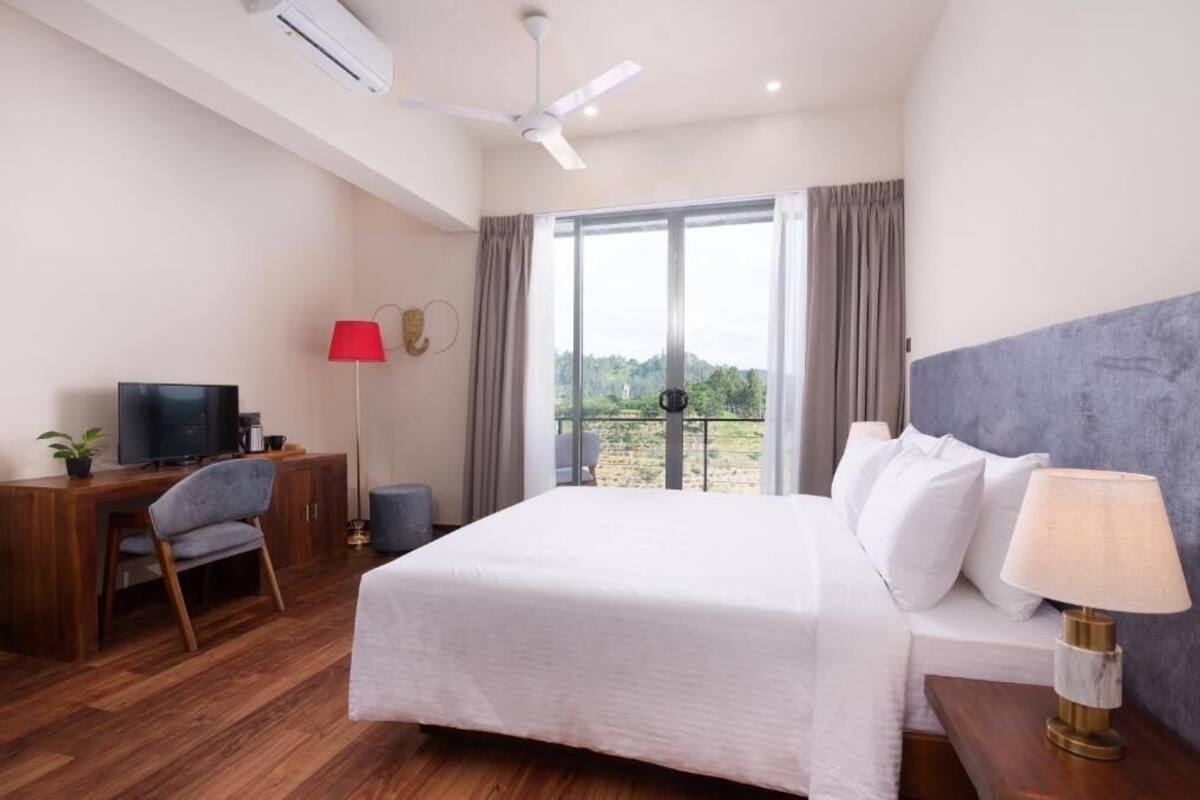 A hotel room with a bed, a desk with a TV, a chair, a lamp, and a view of the outdoors.