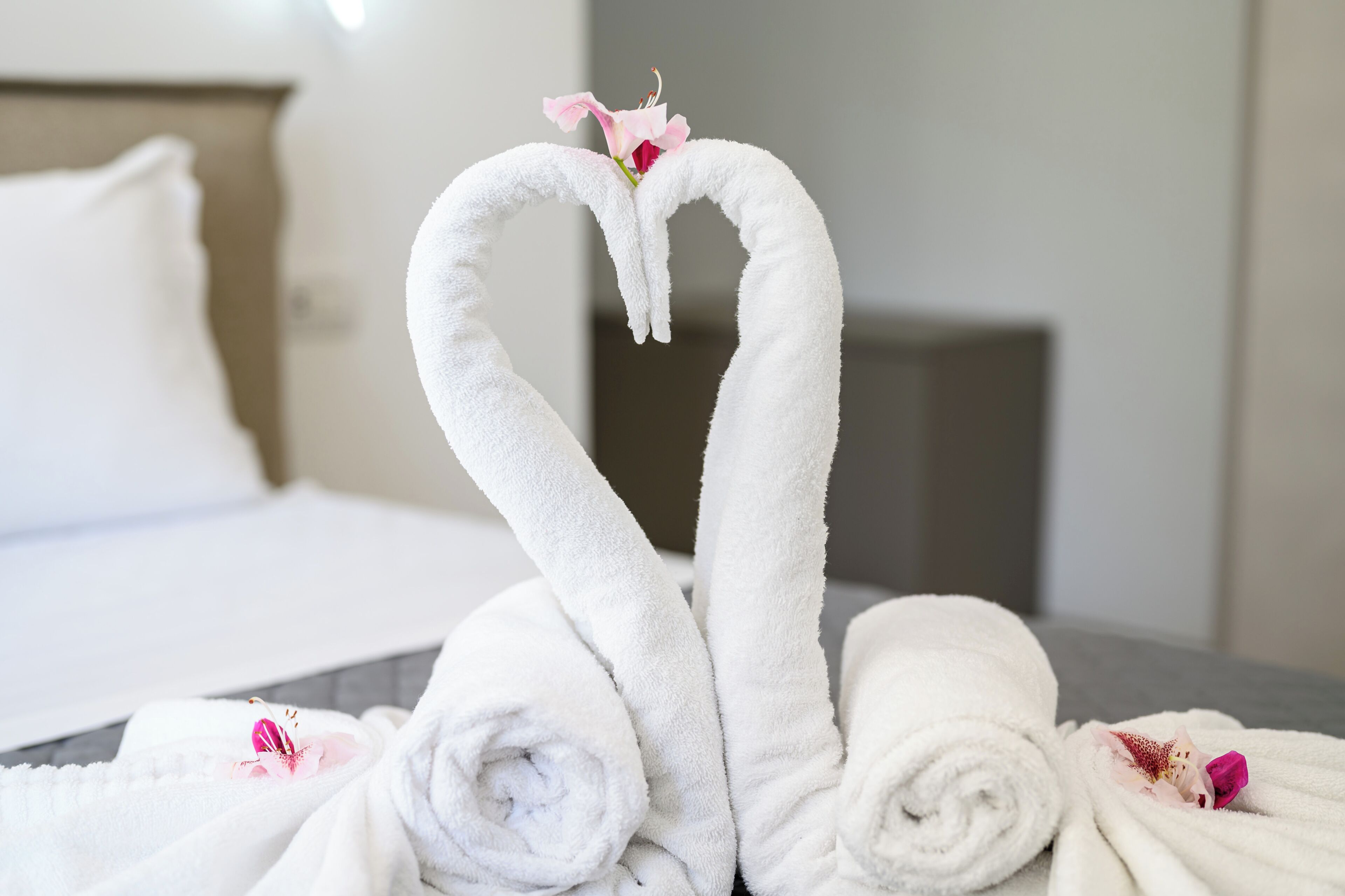 A bed with white towels arranged in a heart shape, a pink flower, and a rolled towel on a hotel room.