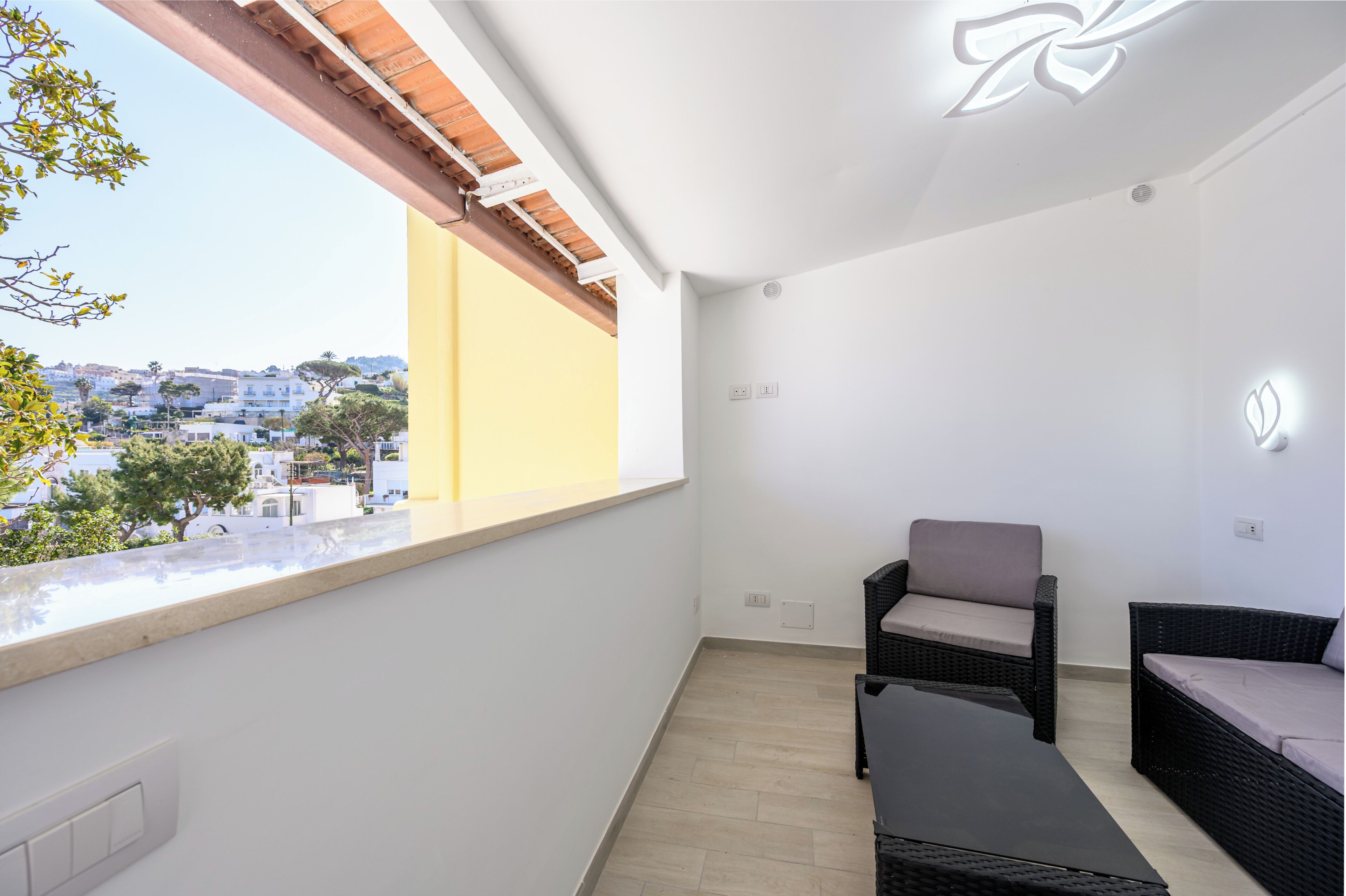 Un balcon donnant sur des immeubles, un canapé en osier, une petite table et un luminaire suspendu au plafond.