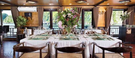 A dining area with tables set for four, each with a vase of flowers, and a view of a natural landscape through large windows.