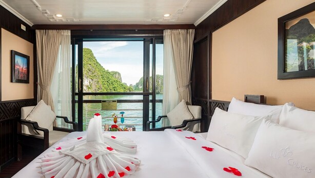 A hotel room with a bed, two chairs, a large window with curtains, and a view of a mountainous landscape. - Lan Ha Bay 4 Star Cruise  (Tuan Chau)