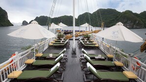 A deck with rows of sun loungers, umbrellas, and tables, overlooking a body of water with hills in the background. - Lan Ha Bay 4 Star Cruise  (Tuan Chau)
