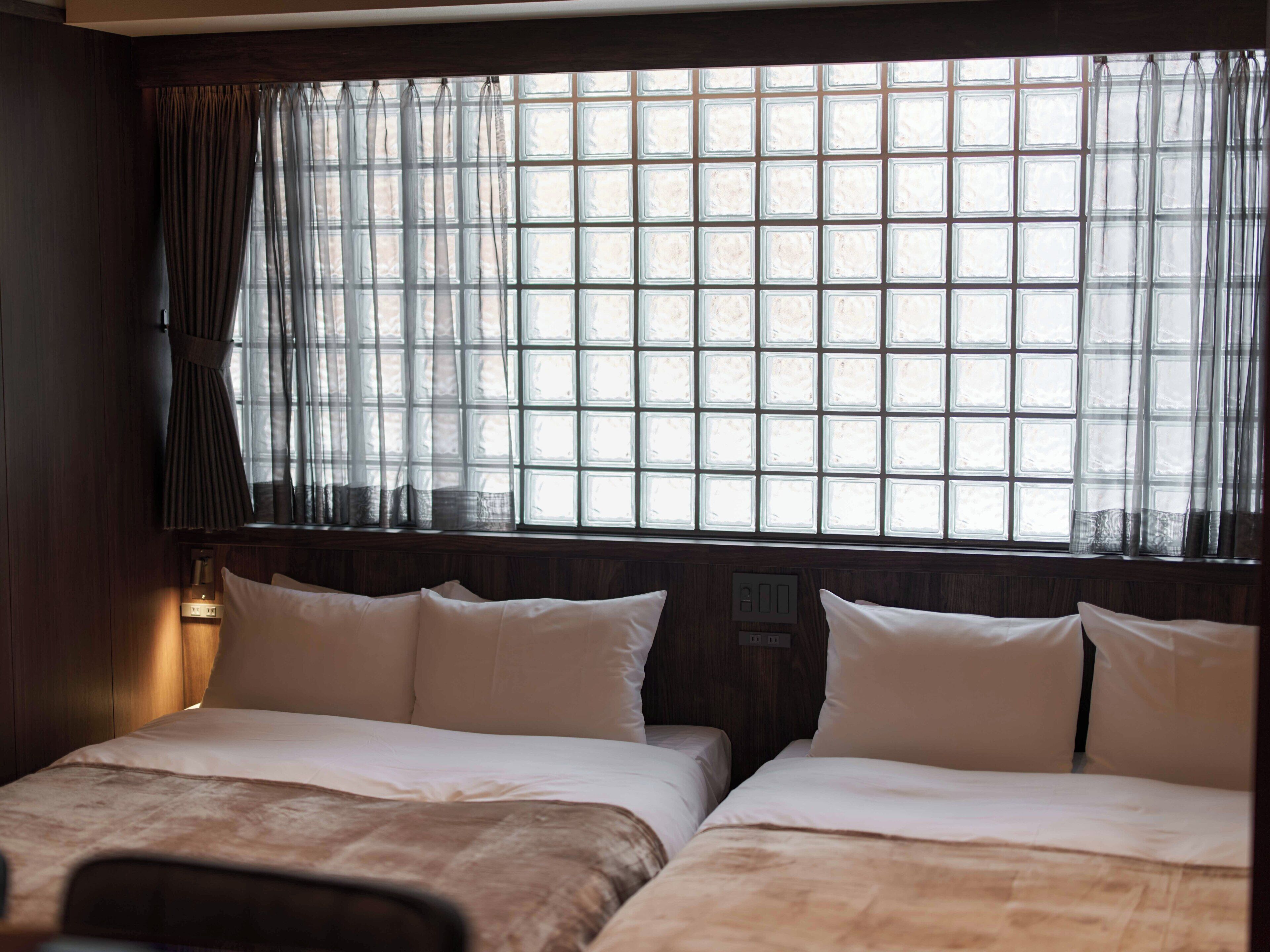 A hotel room with two beds, a window with a grid pattern, and a wall-mounted light.