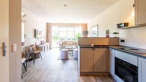Cozinha moderna com forno embutido, área de refeições com mesa e cadeiras, e sala de estar com sofá e mesa de centro.