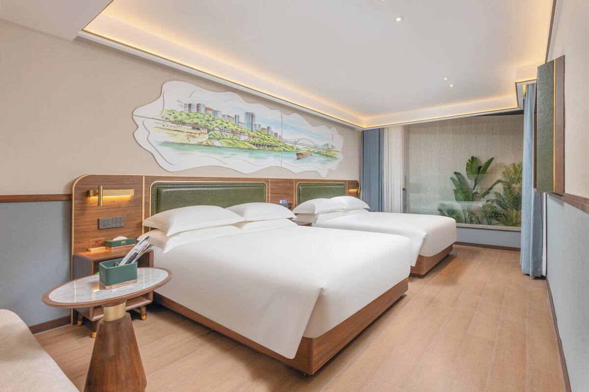 Habitación de hotel con dos camas, una mesita, una mesita de noche, un cuadro colgado en la pared y vistas al verdor a través de una ventana.
