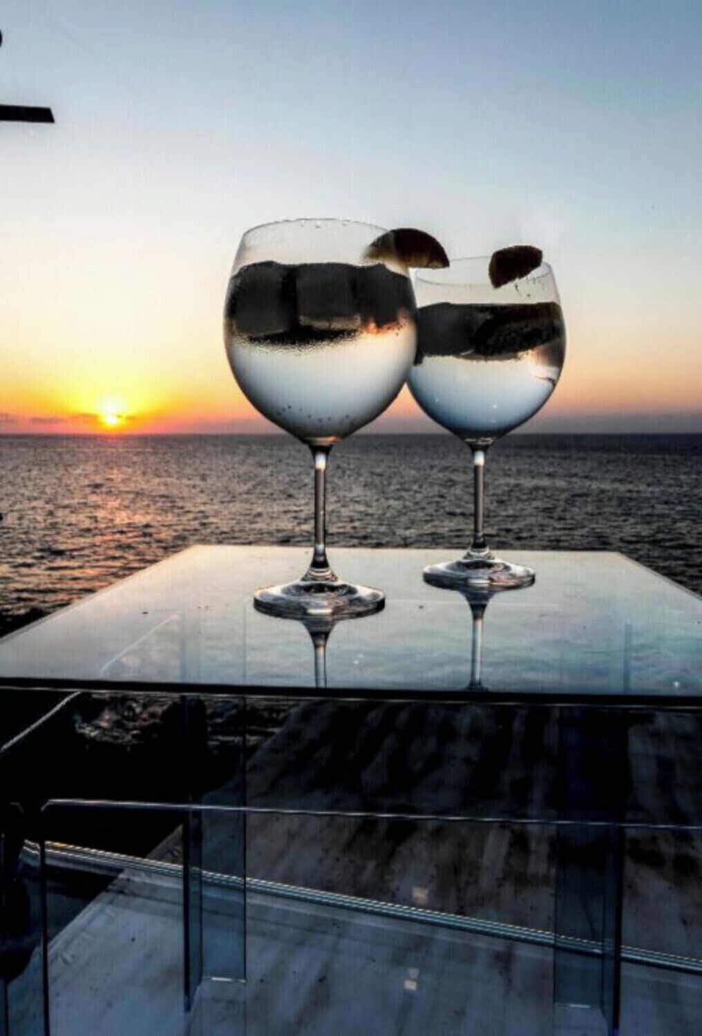 Deux verres à vin sont posés sur une table, offrant une vue sur l’océan au coucher du soleil.