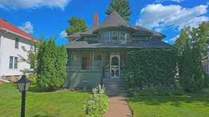 Une maison de style victorien, peinte en vert, avec une véranda qui fait le tour de la maison, une cheminée et un jardin bien entretenu.