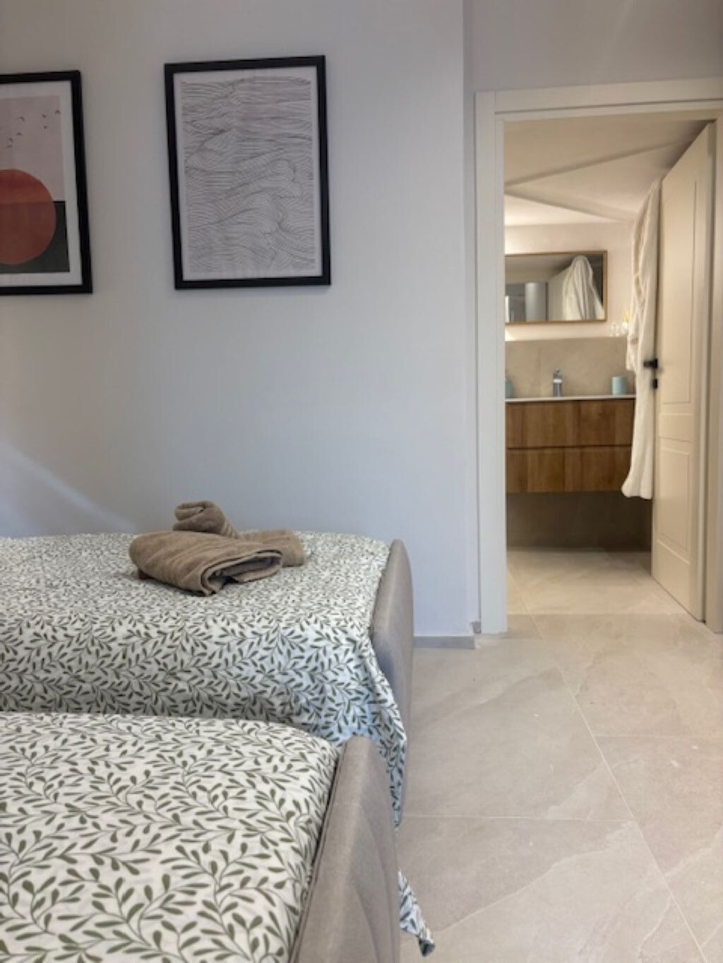 A hotel room with a bed, a bathroom visible through an open door, and two framed artworks on the wall.