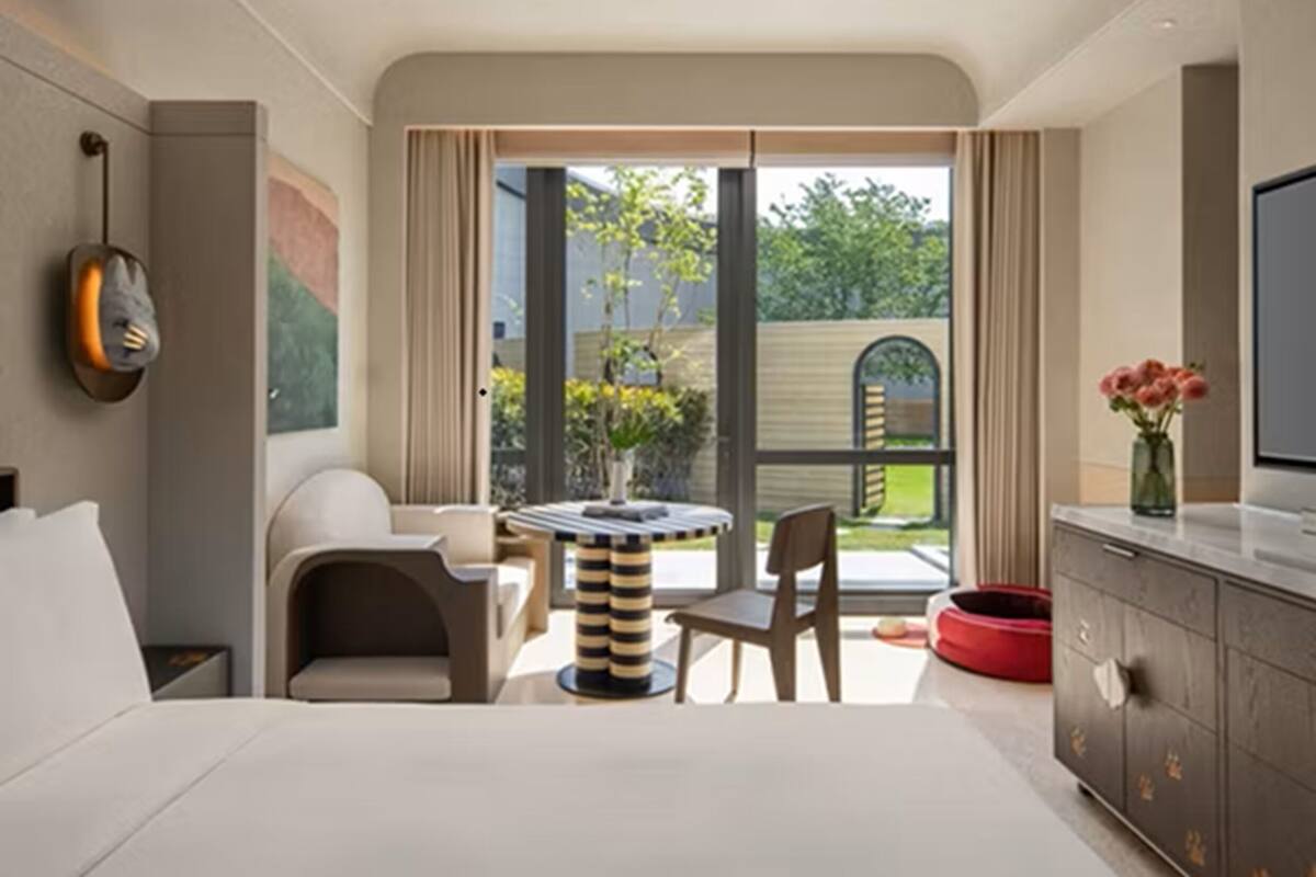 A modern hotel room with a bed, a TV, a dining area with a table and chairs, a red pet bed, and a view of the outdoors.