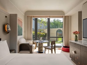 A modern hotel room with a bed, a TV, a dining area with a table and chairs, a red pet bed, and a view of the outdoors. - Kimpton Nine Trees Shanghai by IHG (Shanghai)