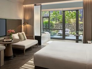 A modern hotel room with a large bed, a seating area with a sofa and armchair, a small table with a lamp, and a view of a garden through large windows.