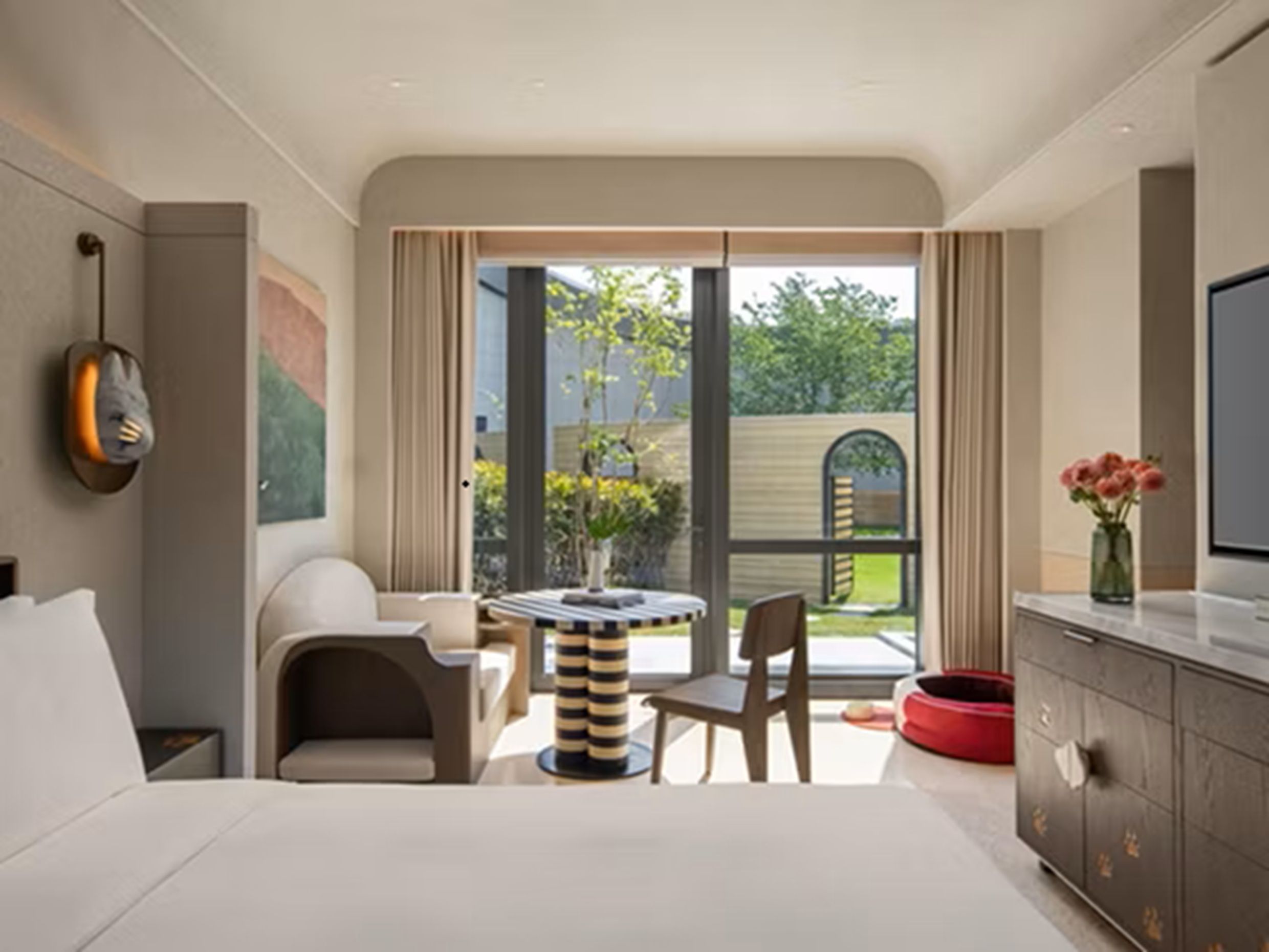 A modern hotel room with a bed, a TV, a dining area with a table and chairs, a red pet bed, and a view of the outdoors.