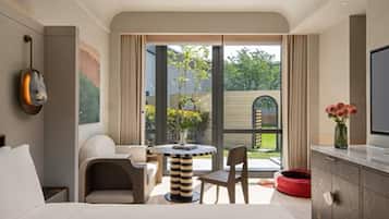 A modern hotel room with a bed, a TV, a dining area with a table and chairs, a red pet bed, and a view of the outdoors.
