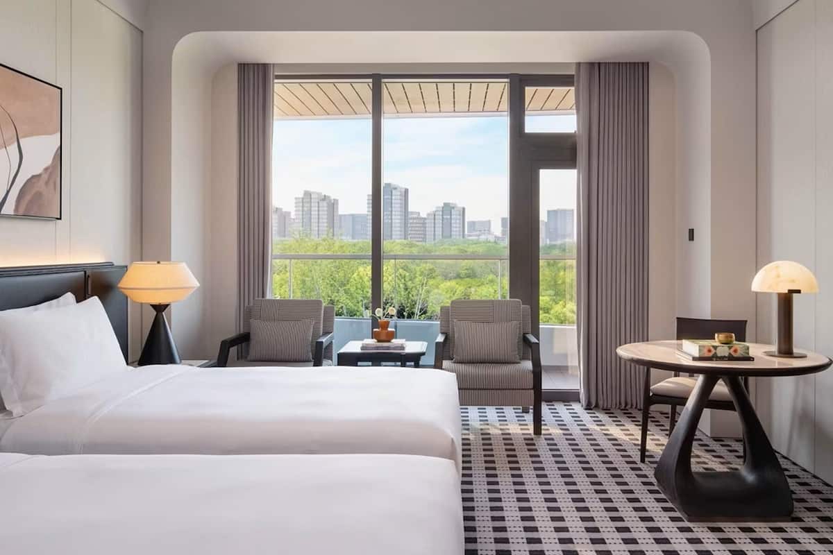A hotel room with a bed, two chairs, a small table, and a view of the cityscape through large windows.