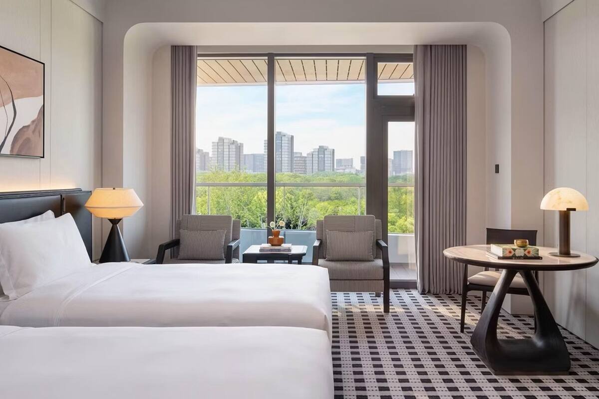 A hotel room with a bed, two chairs, a small table, and a view of the cityscape through large windows.