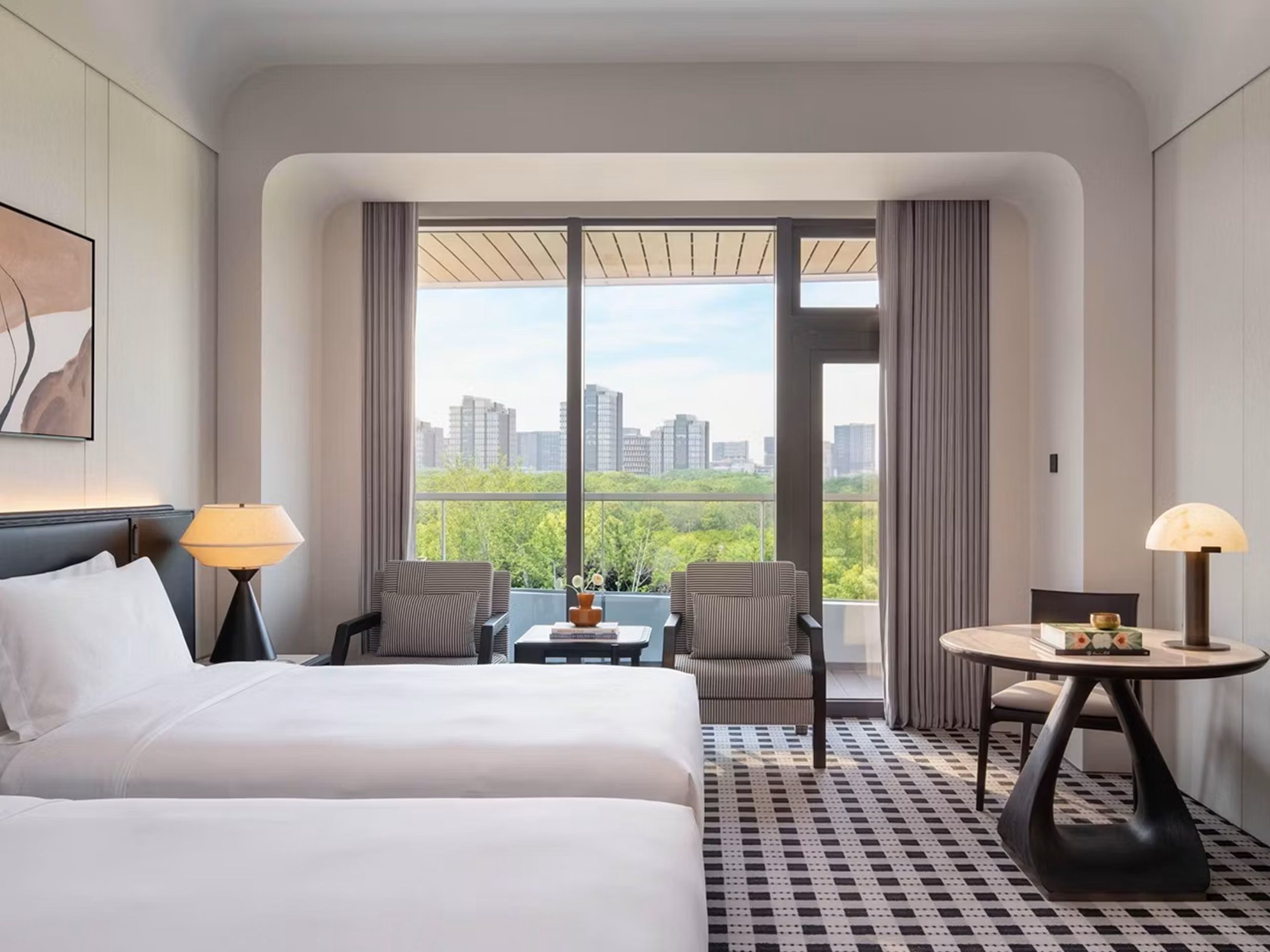 Habitación de hotel con cama, dos sillas, una mesita y vistas a la ciudad a través de amplios ventanales.