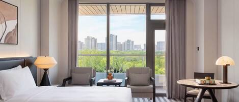 Habitación de hotel con cama, dos sillas, una mesita y vistas a la ciudad a través de amplios ventanales.