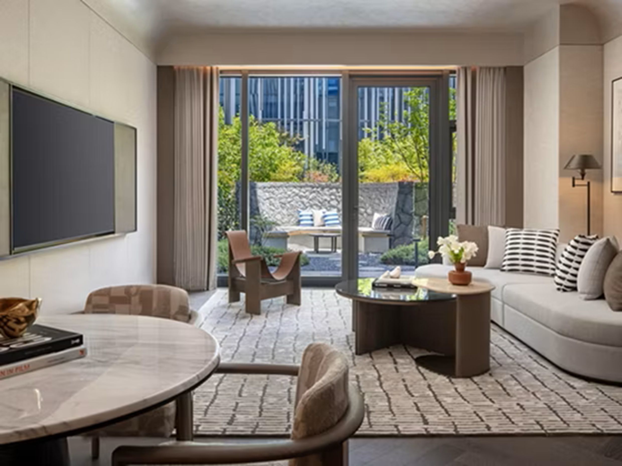 A modern living room with a sofa, coffee table, and chairs, a flat-screen TV, and a view of greenery outside.