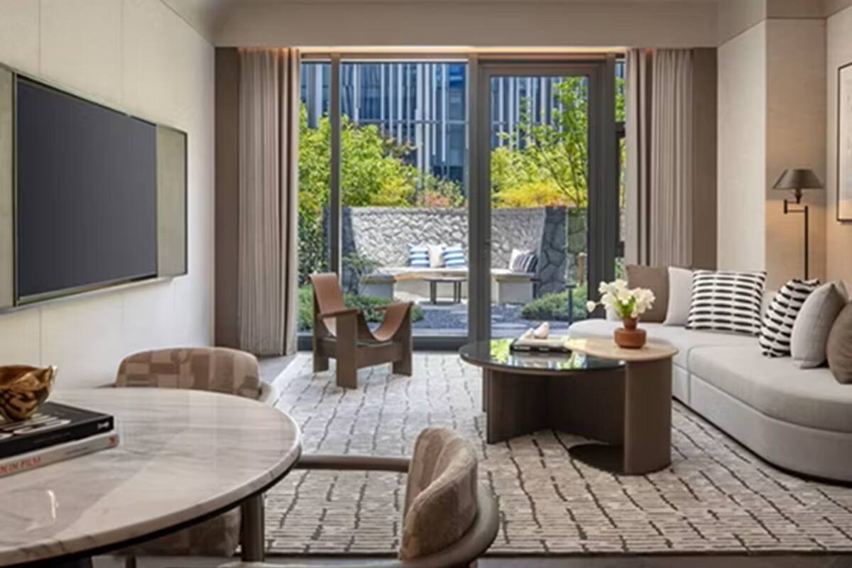 A modern living room with a sofa, coffee table, and chairs, a flat-screen TV, and a view of greenery outside.
