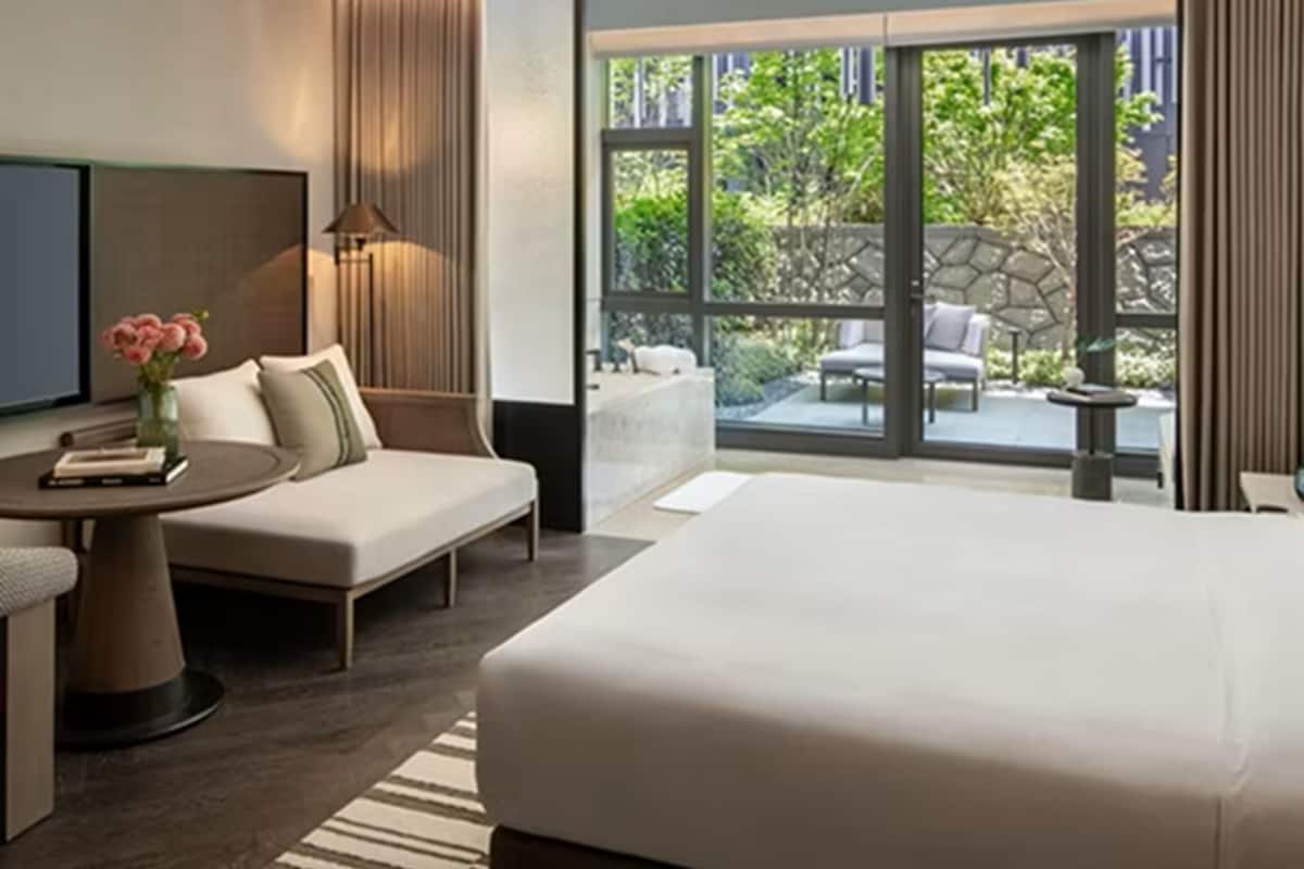 A modern hotel room with a large bed, a seating area with a sofa and armchair, a small table with a lamp, and a view of a garden through large windows.