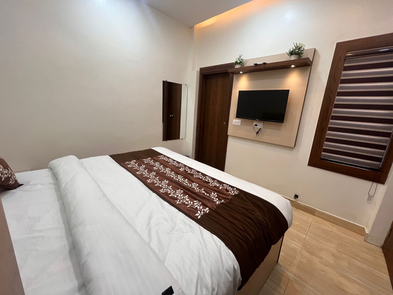 A bedroom with a bed, a television mounted on the wall, and a window with blinds.