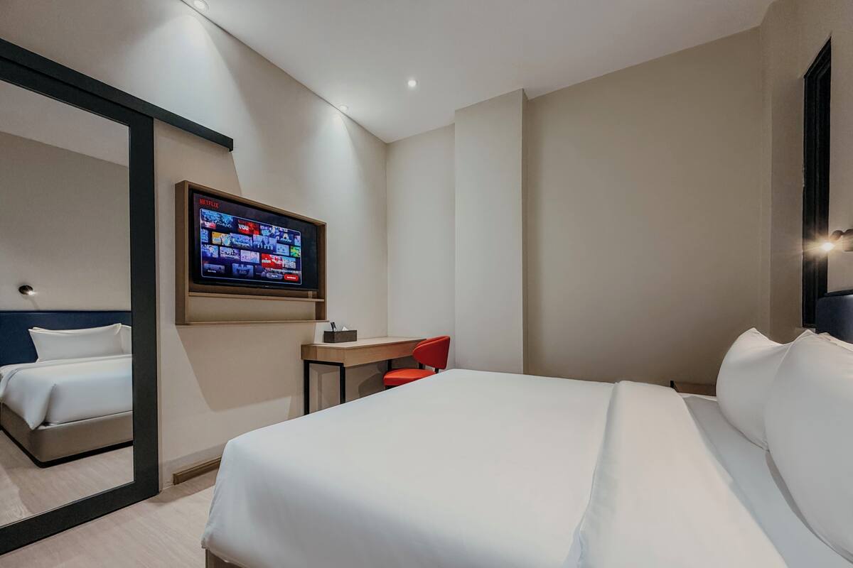 A hotel room with a bed, a desk, a chair, a TV mounted on the wall, and a mirror.