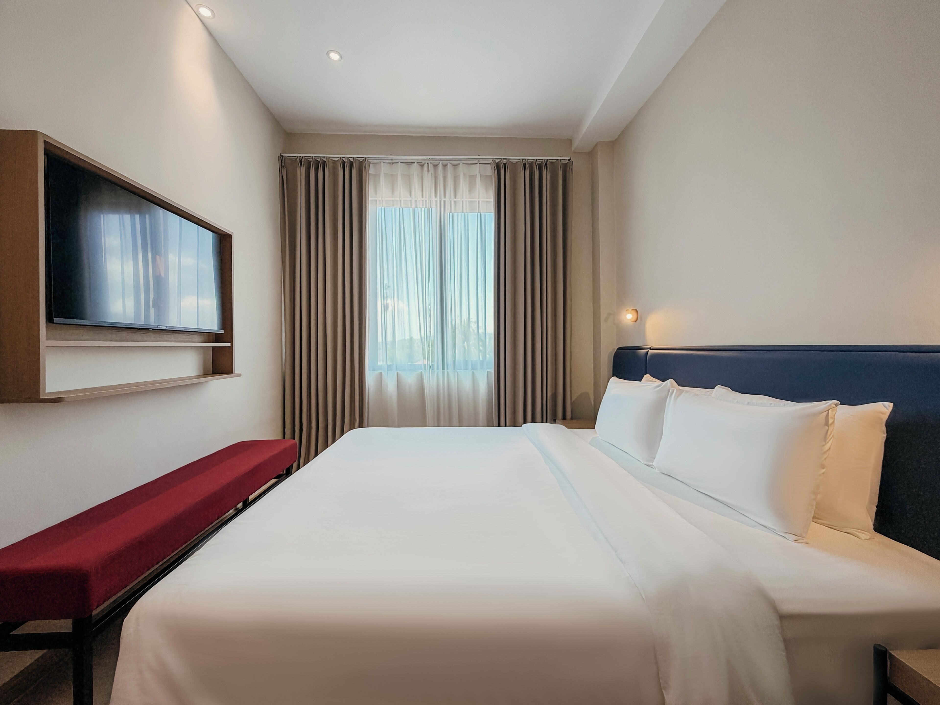 A hotel room with a bed, a TV mounted on the wall, a bench, and a window with curtains.