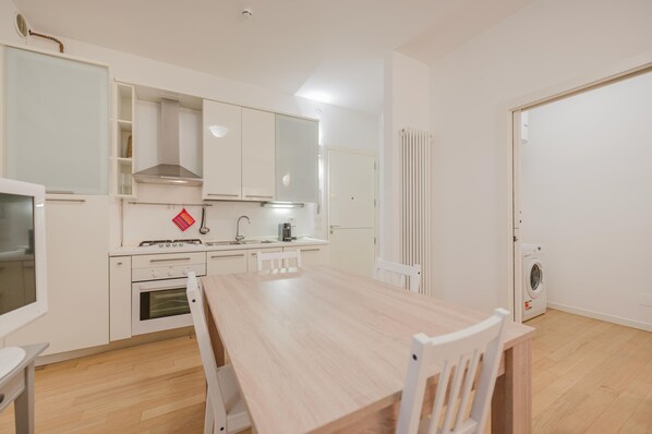 A compact kitchen with white cabinets, a stove, a sink, and a dining table with chairs. - Casa Falco - 15 min dalla stazione (Padova)