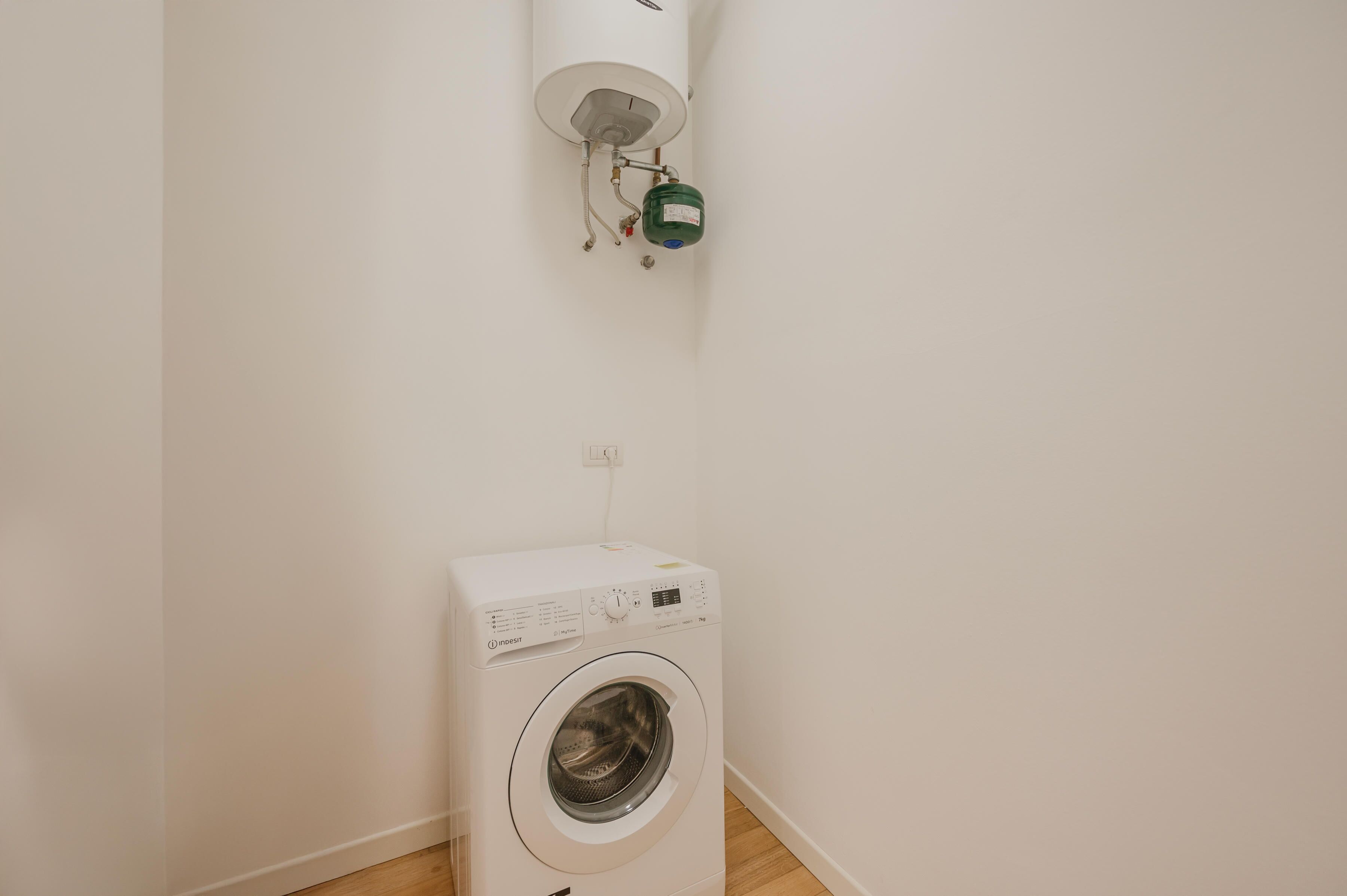 Eine kompakte, weiße Waschmaschine in einem schlichten Raum mit sichtbarer Steckdose und Warmwasserbereiter an der Wand.