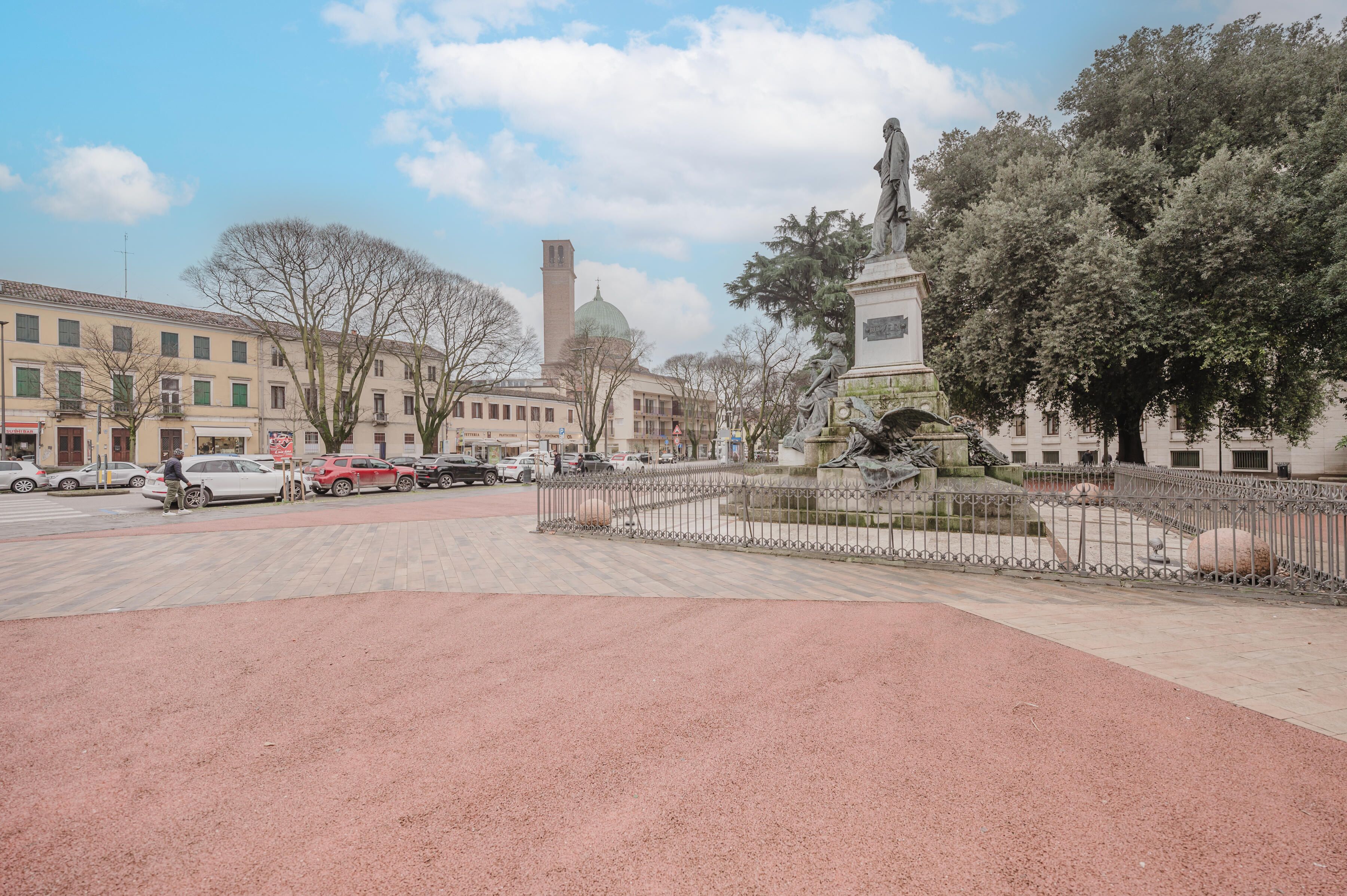 A public square with a statue, a fountain, and a tall tower in the background.