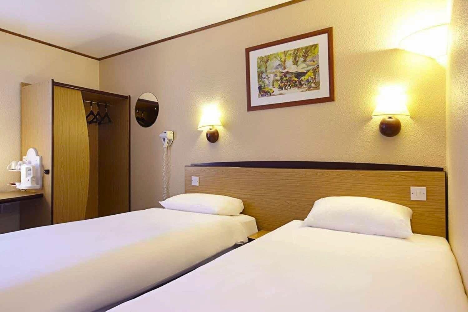 A hotel room with two beds, a bedside table, a mirror, a painting on the wall, and a phone on the wall.