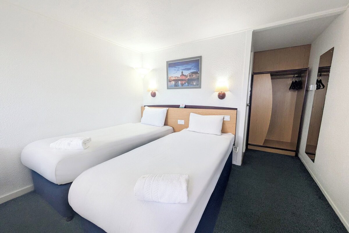 Una habitación de hotel con dos camas individuales, una mesita de noche, una lámpara de pared, un cuadro enmarcado y una puerta que da a un armario.