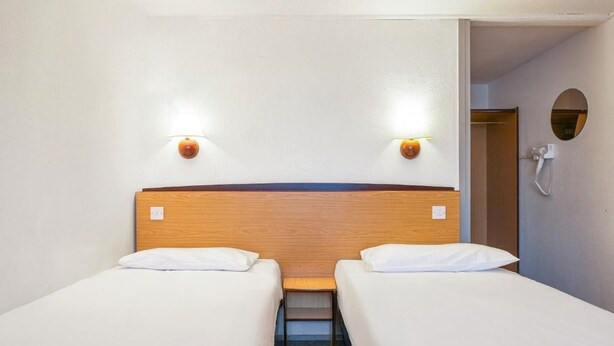 Camera d'albergo con due letti singoli, una testiera in legno, due luci a parete, un tavolino e uno specchio rotondo appeso al muro.