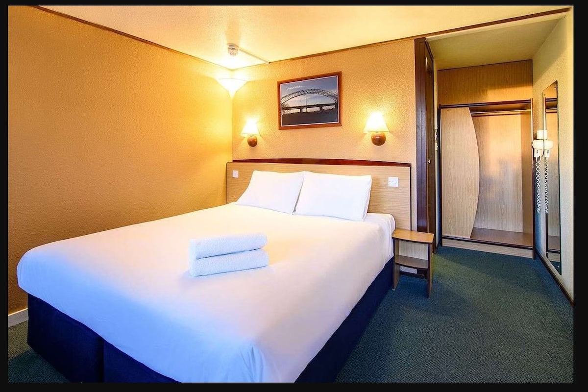 A hotel room with a bed, bedside lamps, a framed picture on the wall, and a door leading to a bathroom.