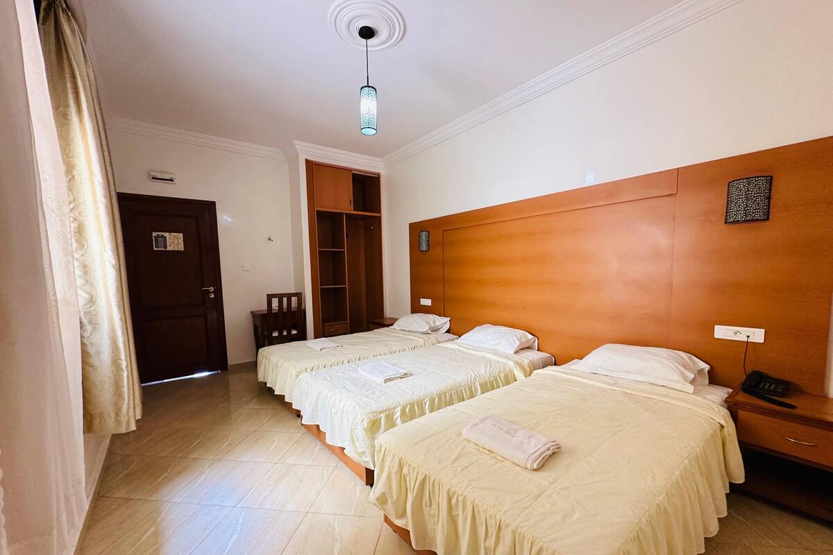 A hotel room with two beds, a wooden headboard, a bedside table with a phone, a wardrobe, and a ceiling light fixture.