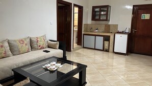 Una sala de estar con un sofá, una mesa de centro de vidrio, una zona de cocina con armarios y un refrigerador, y una puerta que da a otra habitación.