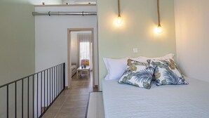 Un dormitorio con cama, almohadas, una mesita de noche, una ventana y una puerta que da a otra habitación.