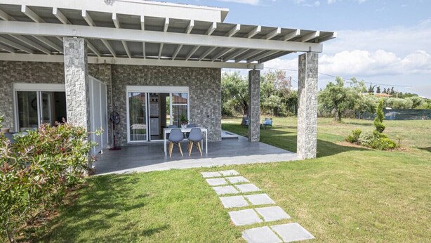 Una casa moderna con fachada de piedra, un porche cubierto con mesa y sillas, y un camino de piedra que lleva a la entrada.