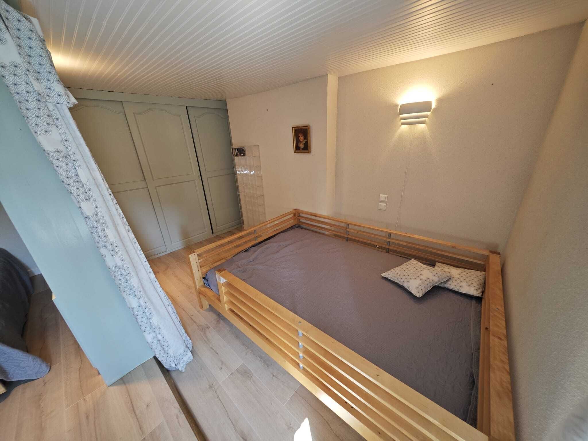 A bedroom with a wooden bed frame, a light-colored carpet, a wall-mounted light fixture, and a door with a curtain.