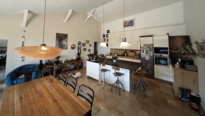 Una cocina moderna con armarios blancos, una isla central, luces colgantes y una zona de comedor con mesa y sillas de madera.