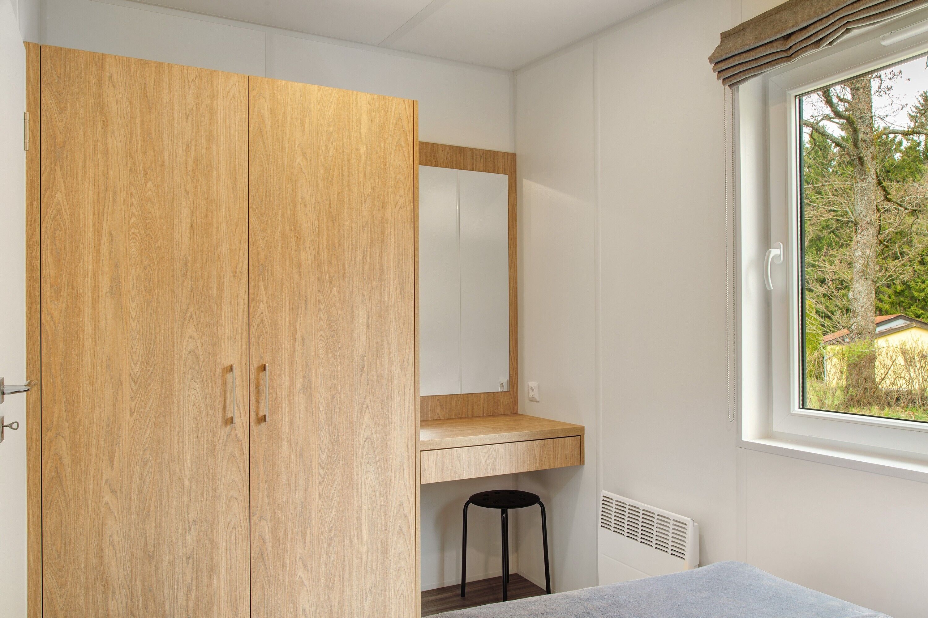 A bedroom with a wooden wardrobe, a mirror, a window with a view of trees, and a radiator.