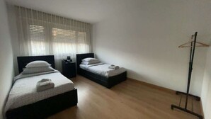 Een slaapkamer met twee bedden, een nachtkastje, een kapstok en een raam met jaloezieën.