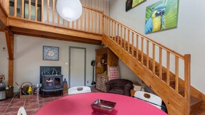A spacious living area with a wooden staircase, a round table, a fireplace, and a parrot painting. - Holiday Home 'Aux Agapanthes', Shared Pool, Private Terrace, Landscaped Park, Free Wi-Fi (Aumagne)