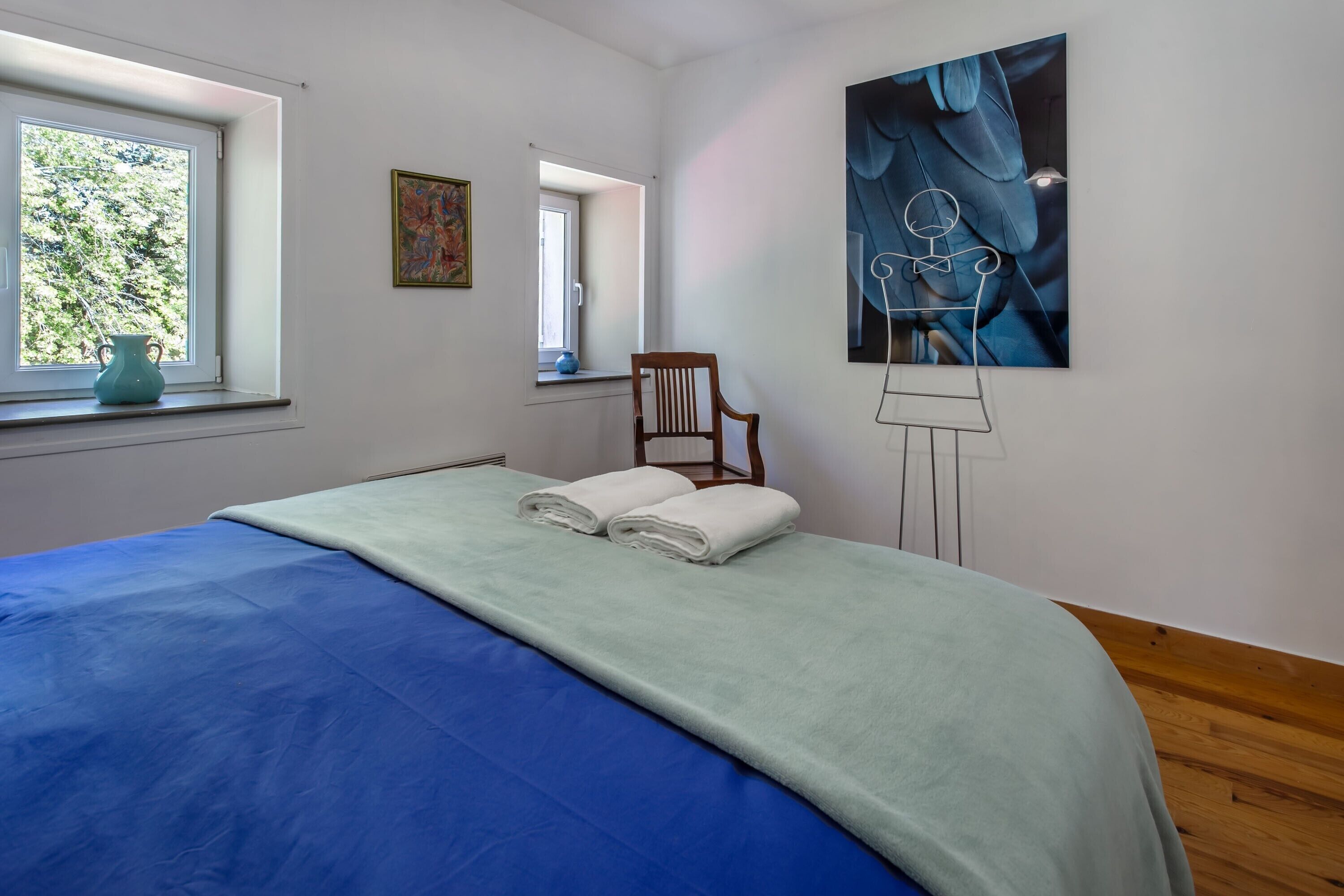 Ein Zimmer mit einem Bett, einem Fenster, einem Stuhl und einem Bild an der Wand.