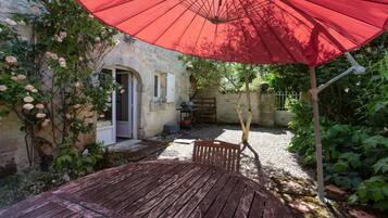 Un espace extérieur aménagé dans un style rustique, avec une table et un banc en bois, un parasol rouge et un mur de pierres sur lequel grimpent des rosiers.