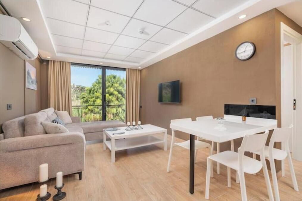 A modern living room with a sofa, a coffee table, a dining table with chairs, a TV, and a clock on the wall.