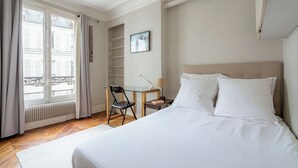 A hotel room with a bed, a desk, a chair, a window with curtains, and a radiator. - Parisian Muse, by Veeve (Paris)