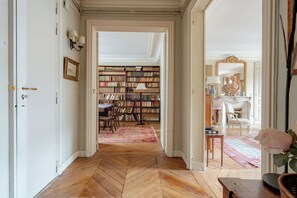 Un corridoio con pavimento a spina di pesce che conduce a una stanza con libreria, uno specchio e una statua.