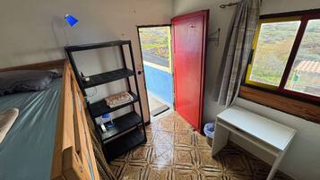 Una habitación pequeña con una cama, una estantería, un escritorio, una puerta roja y una ventana.